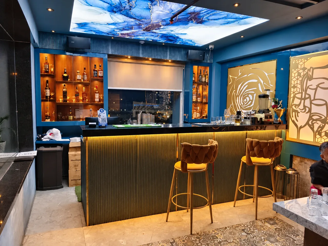 Illuminated indoor bar with gold stools, bottle shelves and a blue ceiling panel