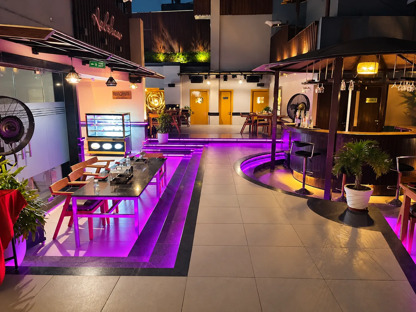 Rooftop walkway between dining tables and a circular bar with purple lighting