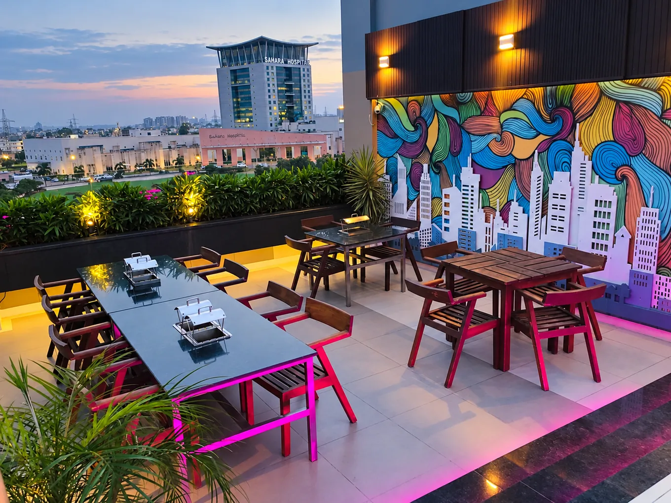 Wooden rooftop dining tables beside a colourful skyline mural at dusk